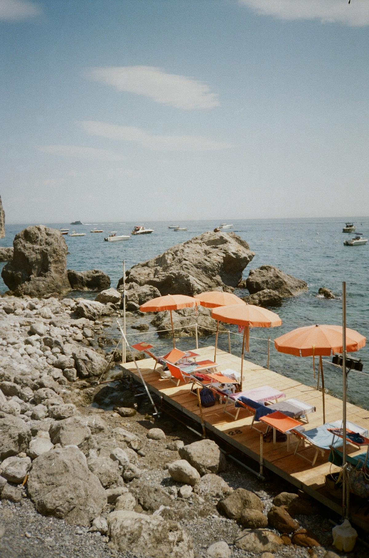 Orange umbrellas and striped chaise longues lined up on a wooden boardwalk at an Italian bathing establishment, with a rocky shore and Mediterranean sea behind