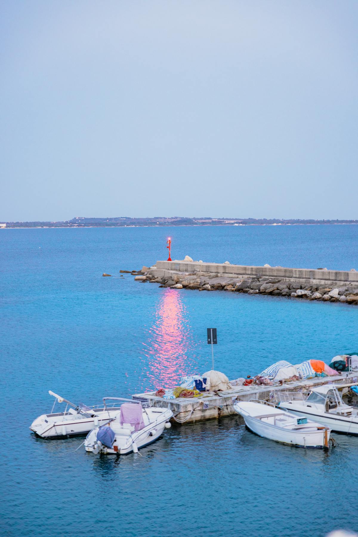 Small moored boats tied alongside a stone quay at a calm harbour entrance, with a navigation light at the jetty