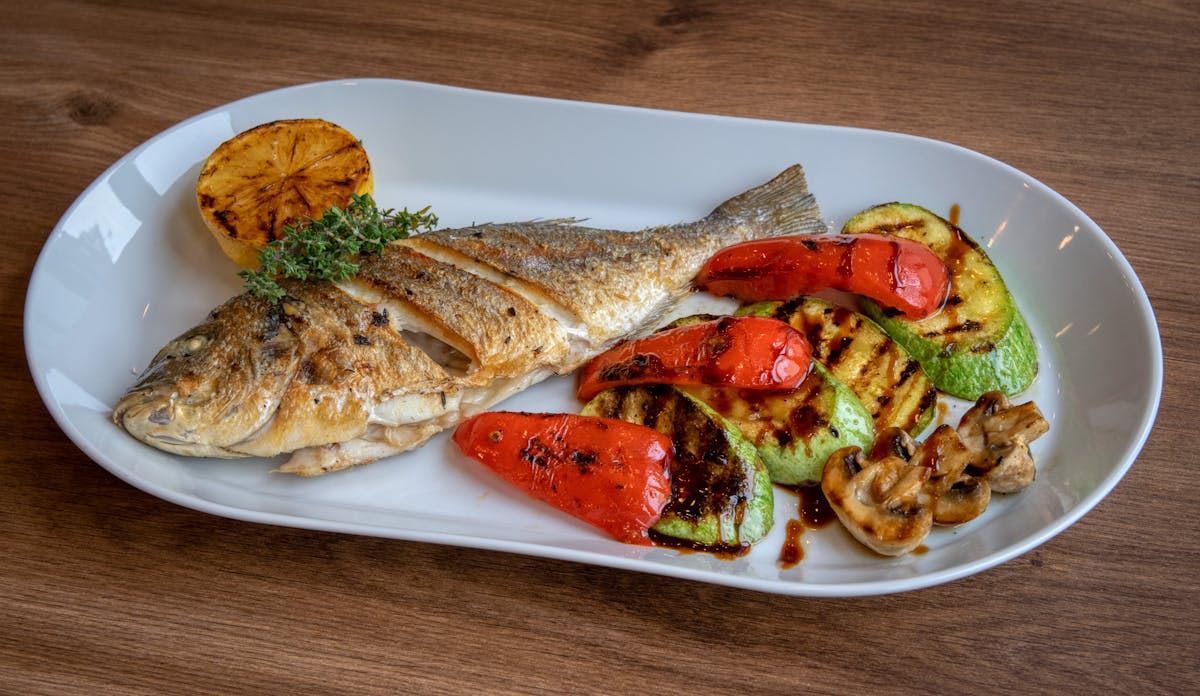 Whole grilled sea bass plated with charred lemon and grilled Mediterranean vegetables on a white oval dish
