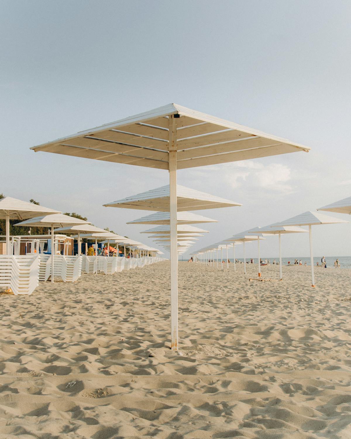 Rows of square modern beach umbrellas on raked sand, with slatted wood structures behind in late afternoon light