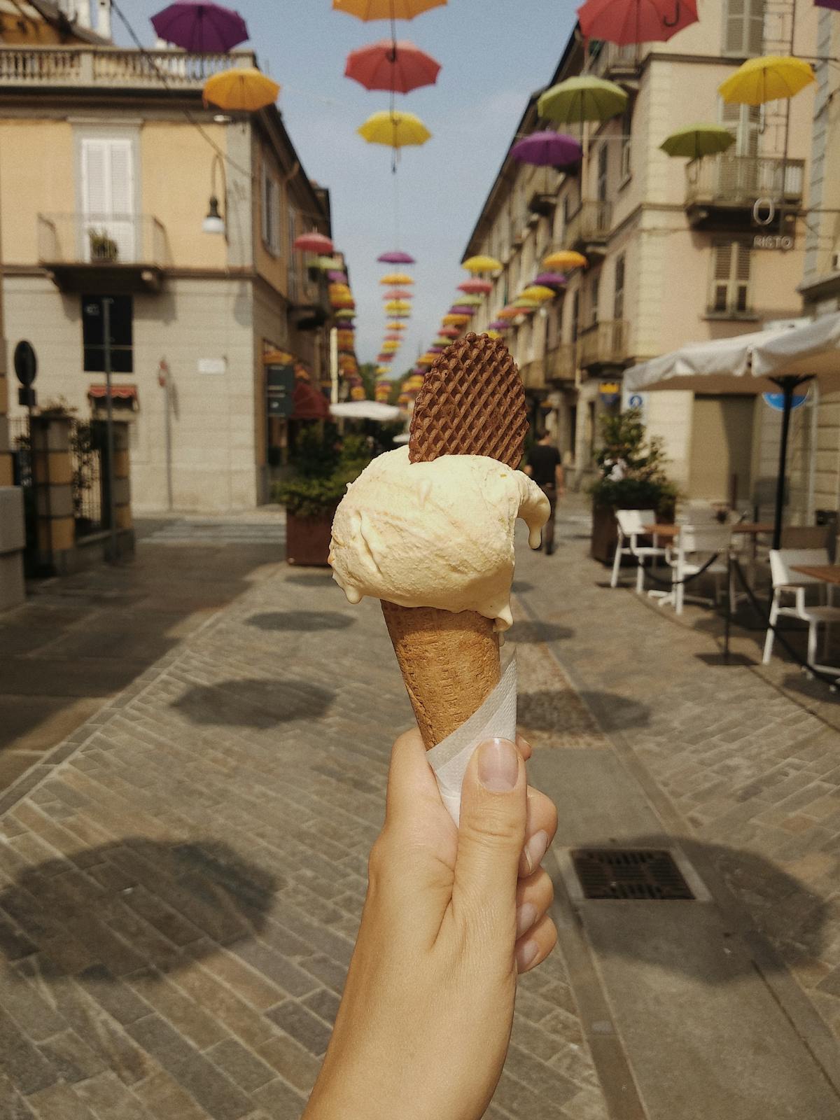 Hand holding an ice-cream cone with a cream-coloured gelato scoop and a wafer, in a pedestrian Italian street hung with rows of coloured parasols overhead