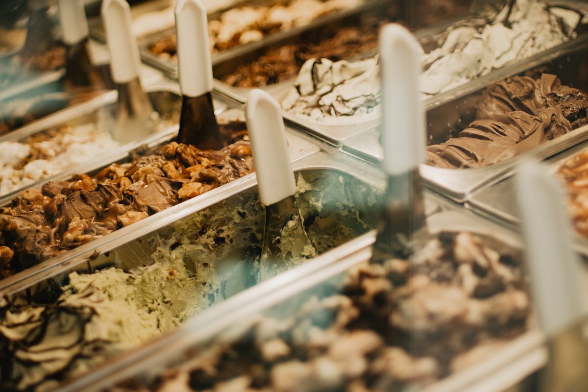 Close-up of multiple open gelato tubs in a chilled display case, showing pistachio, chocolate, cream and sorbetto textures with metal serving paddles upright in each tub