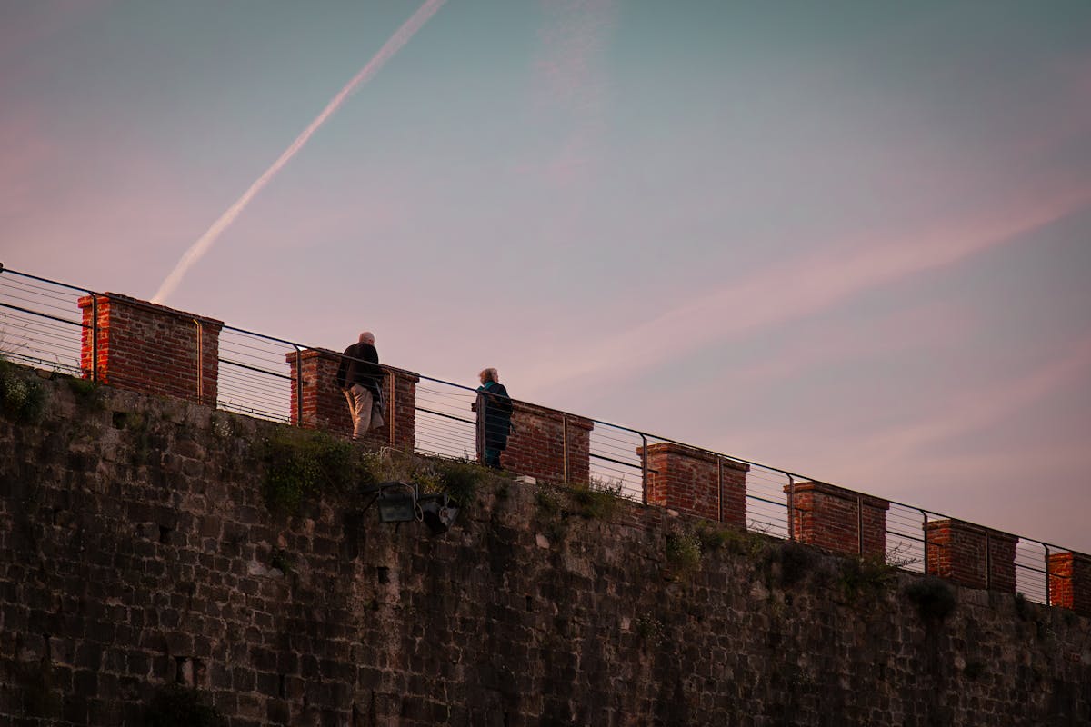 Two figures walking on the raised brick rampart of the Lucca city walls at dusk, pink sky above