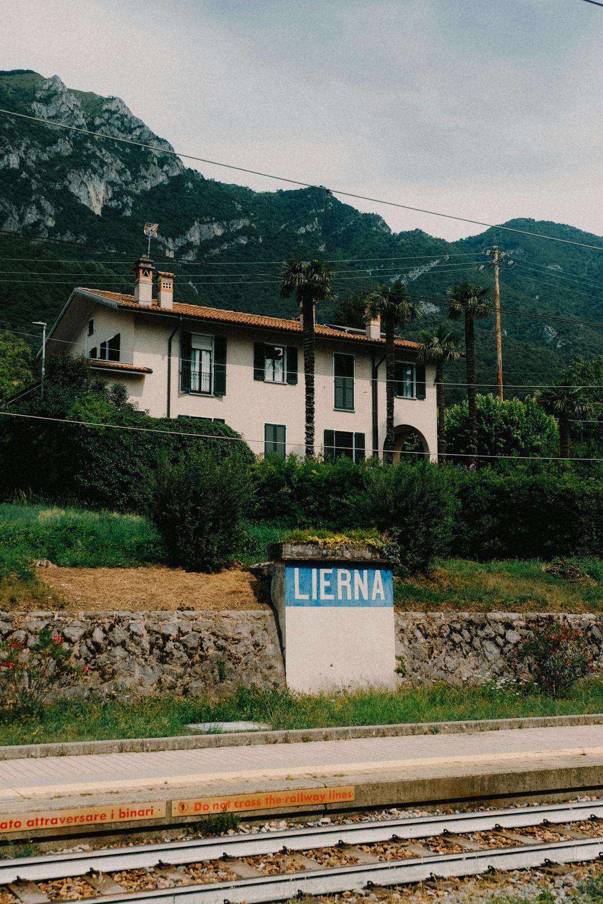 Italian regional railway station building beside the tracks, with palm trees, mountains behind, and a painted station-name sign